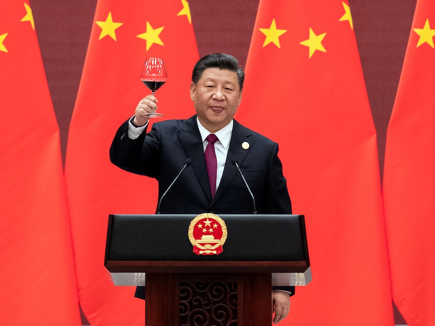 Xi Jinping raises his glass and proposes a toast while standing behind a lecturn in front of Chinese flags.