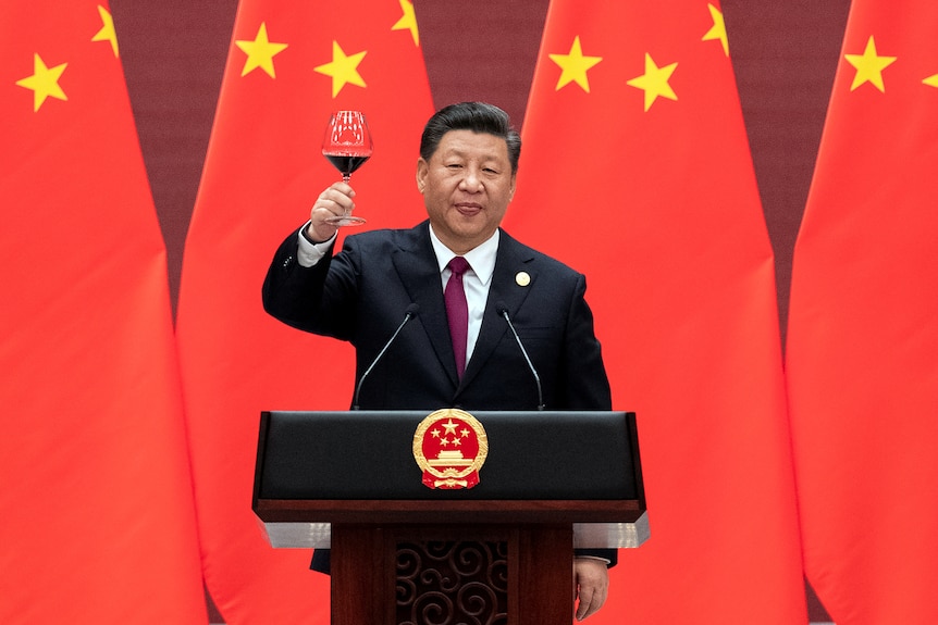 Xi Jinping raises his glass and proposes a toast while standing behind a lecturn in front of Chinese flags.