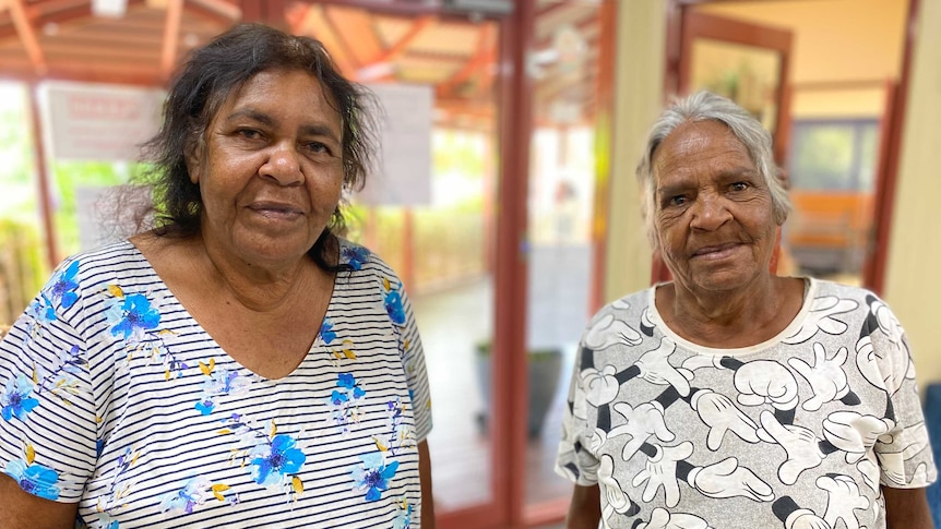Two Indigenous women wearing patterned shirts stand together inside.