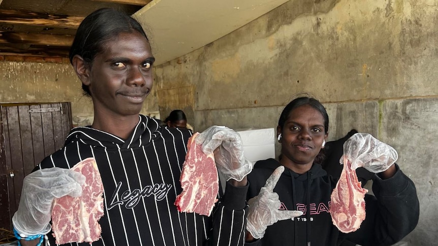 Two young Indigenous women wearing aprons stand together, learning to butcher meat.