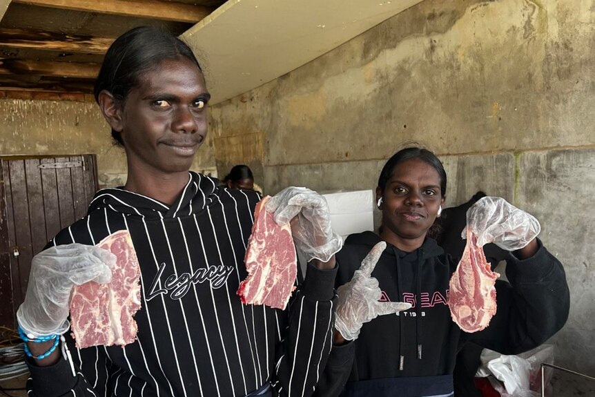 Two young Indigenous women wearing aprons stand together, learning to butcher meat.