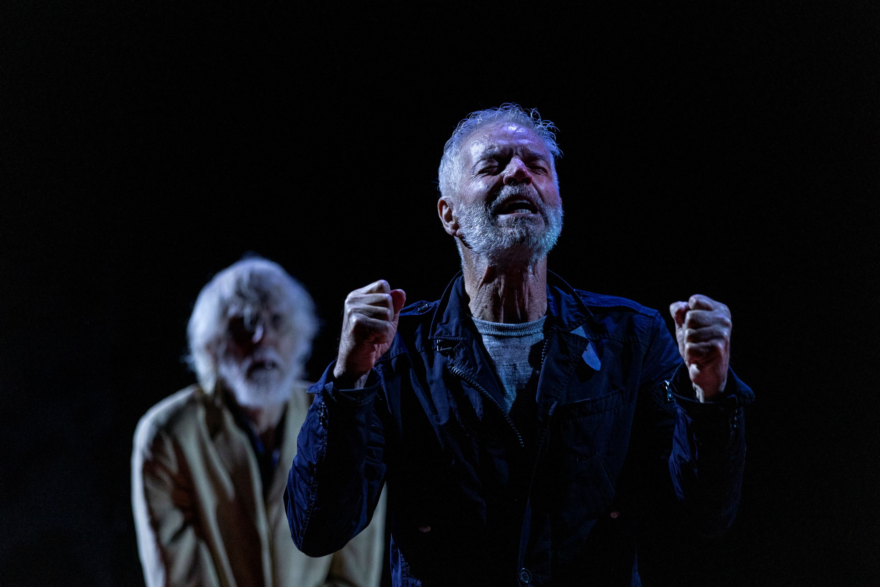 En el escenario, envuelto en sombras, Colin Friels, de 73 años, parece dolido, con los puños cerrados en el aire. Peter Carroll está detrás de él.