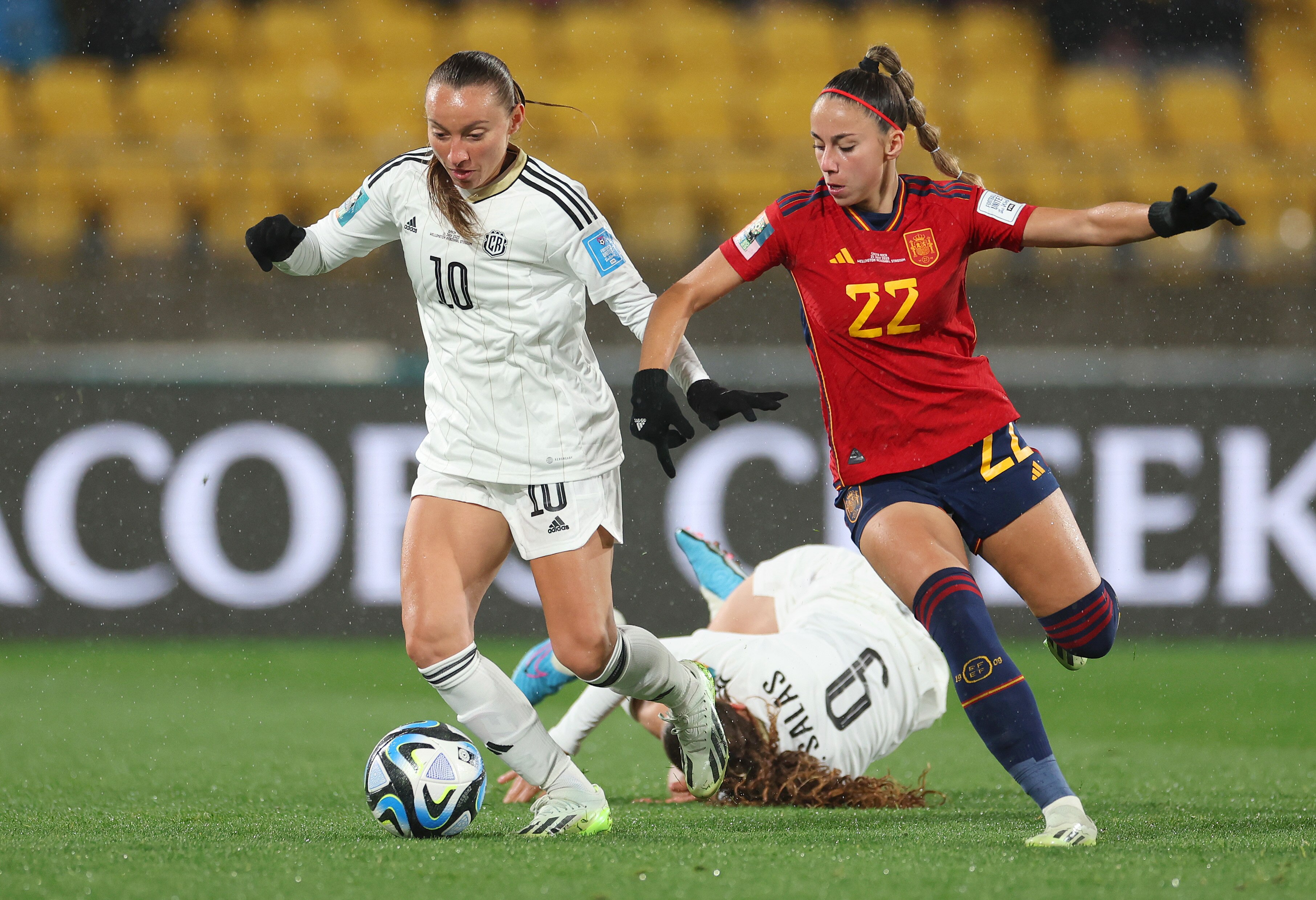 Two opposing players fight for the ball while another player falls to the pitch behind them.