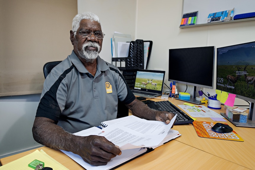 An Aboriginal man in a grey polo shirt sits at a desk holding papers