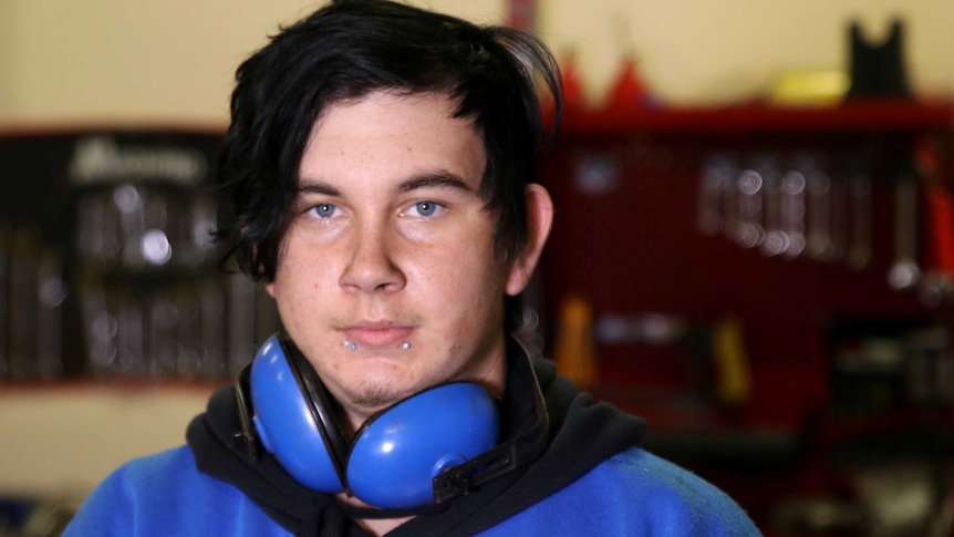 A teenage boy with facial piercings and wearing a pair of ear muffs around his neck looks at the camera.