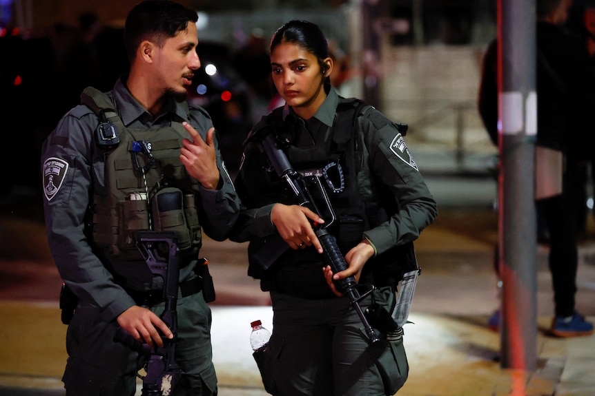 A young man and women who are armed members of the Israel security forces guard the scene of a shooting attack at night.