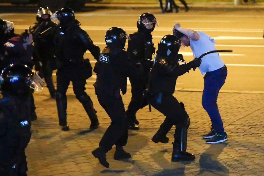 Police wearing riot armour swing a rubber truncheon into the left flank of a man who has raised his hands above his head.