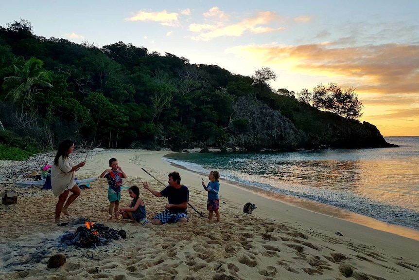 a family sit on a beach next to campfire with a sunset and jungle in the background