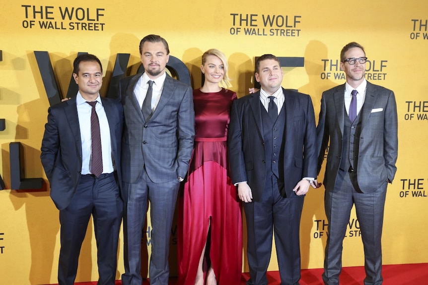The two producers and three cast members stand on red carpet in front of yellow Wolf of Wall Street wall