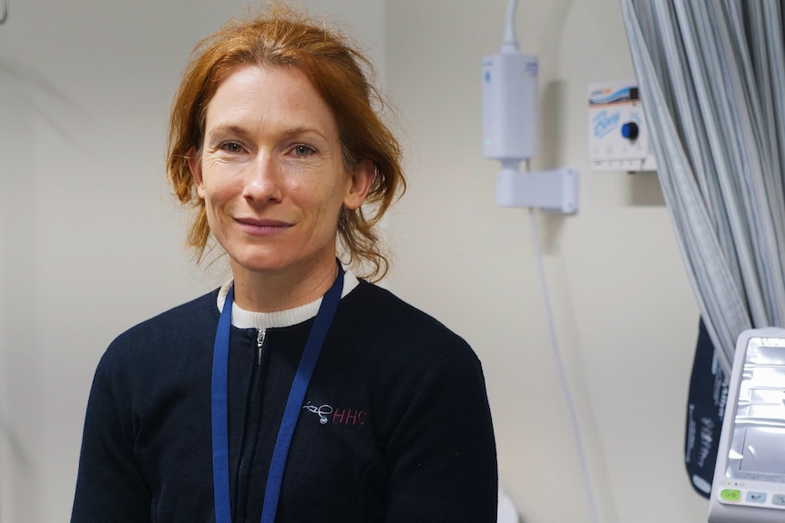 A woman with ginger hair, wearing a dark top and a lanyard, stands in what appears to be a medical clinic.