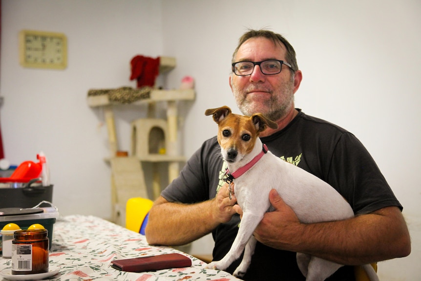 A man holds a small white and brown dog in his arms while sitting at a table in a room