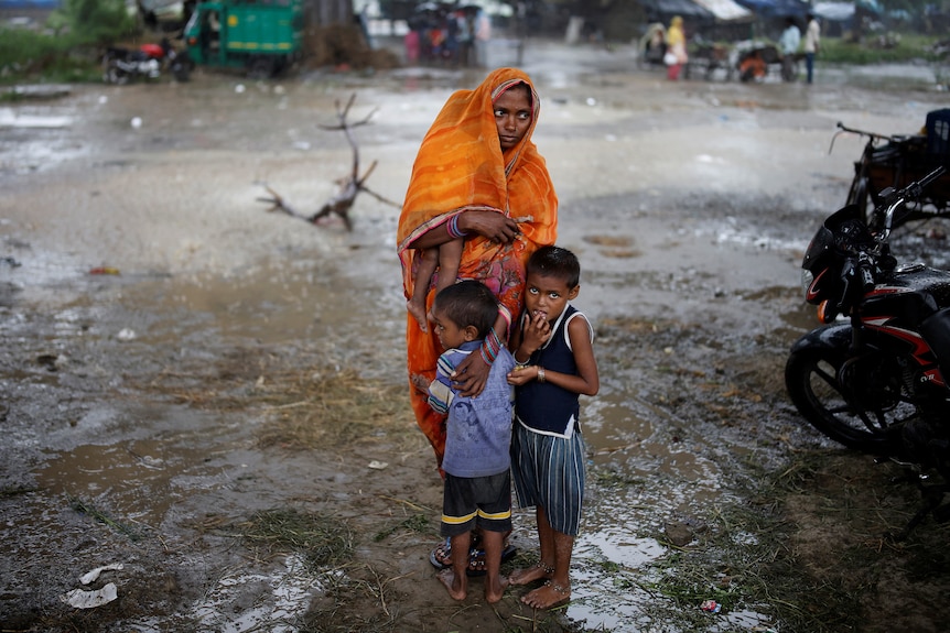 A mother stands wrapped in an orange sari, holding a young child with two standing at her feet. 