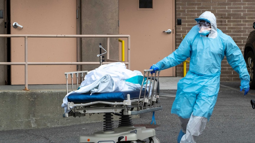 A health worker, head to toe in protective equipment, runs a body on a stretcher through a car park