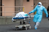A health worker, head to toe in protective equipment, runs a body on a stretcher through a car park
