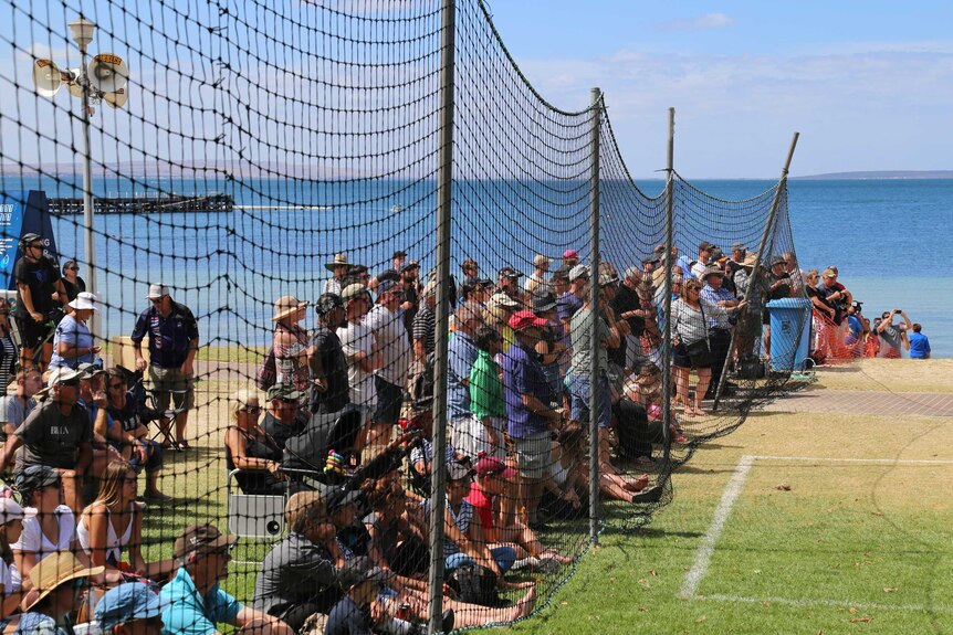 A hundred or so people stand behind a five-metre high black net.