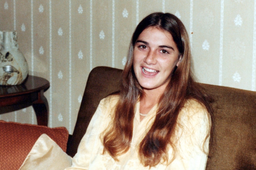 A young woman with long brown hair sits on a couch, smiling.