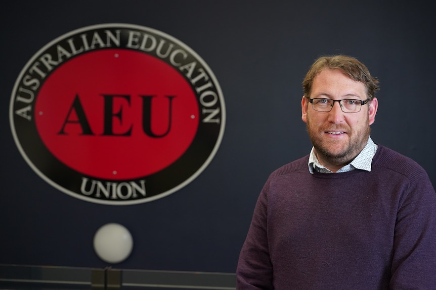 A middle-aged man in glasses on the right of the frame next to a large AEU sign.