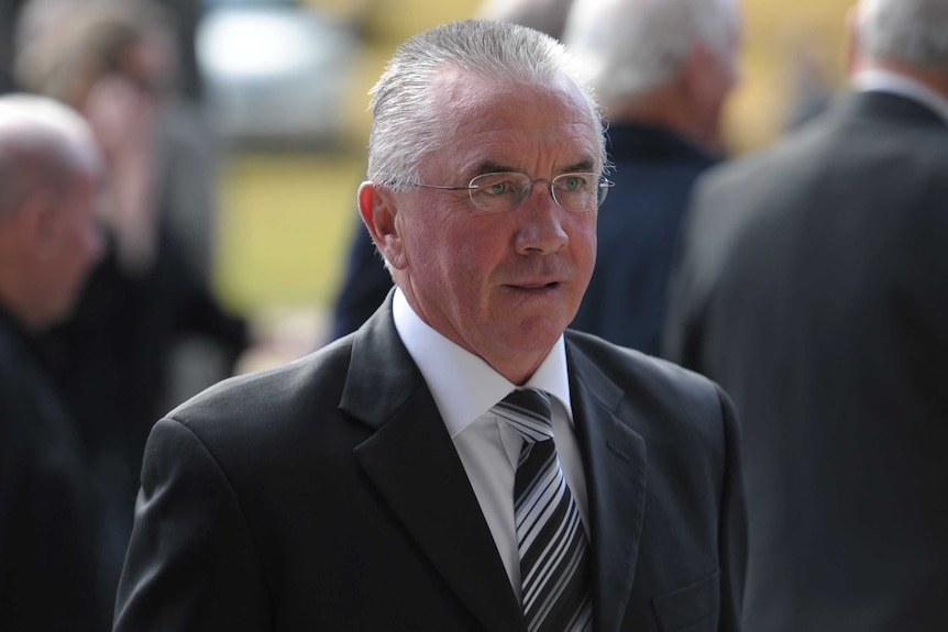 A former AFL player and coach stands in a suit at a funeral service at the MCG.