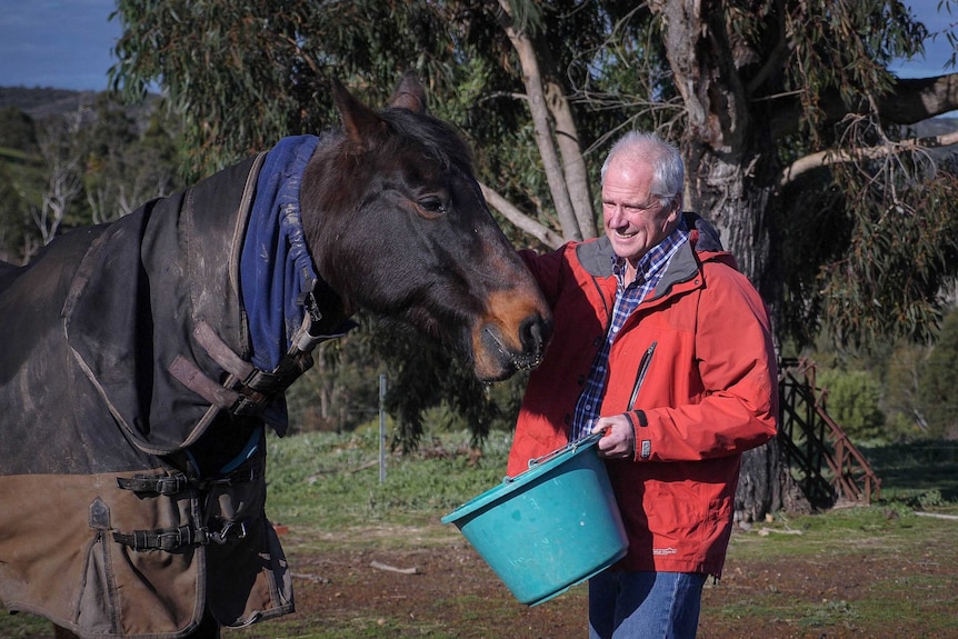 A man with white hair wears a red jacket and is holding a green bucket that he's using to feed a horse.