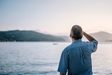 Man seen from behind with grey hair and blue shirt. He faces towards a vast expanse of blurred ocean.
