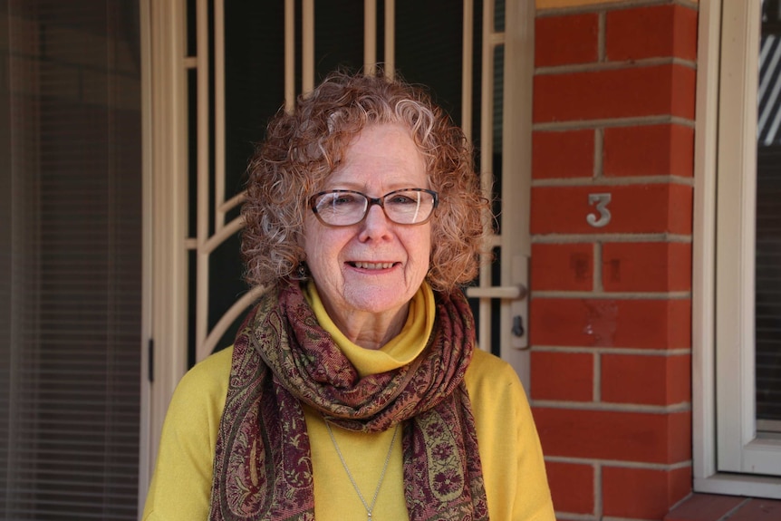 A woman wearing glasses and a patterned maroon scarf smiles while standing before the front door of a brick house.