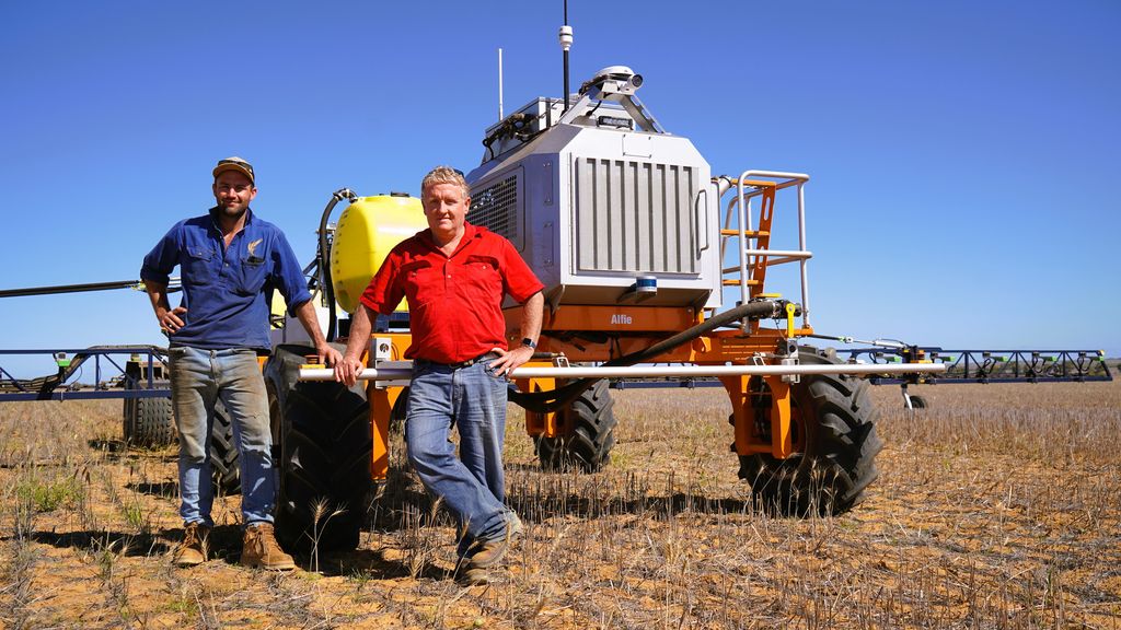 An older and young man stand next to tractor-looking robot in a paddock with stubble, blue skies.