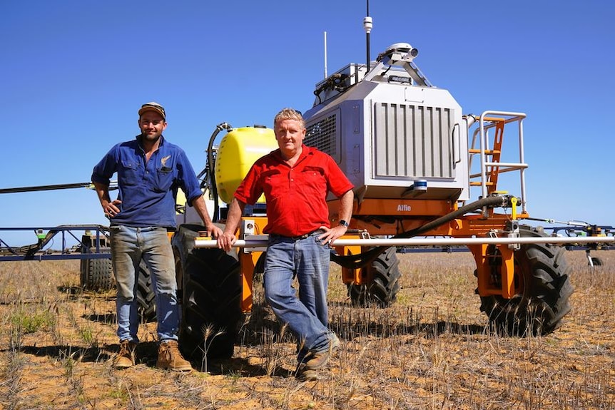An older and young man stand next to tractor-looking robot in a paddock with stubble, blue skies.