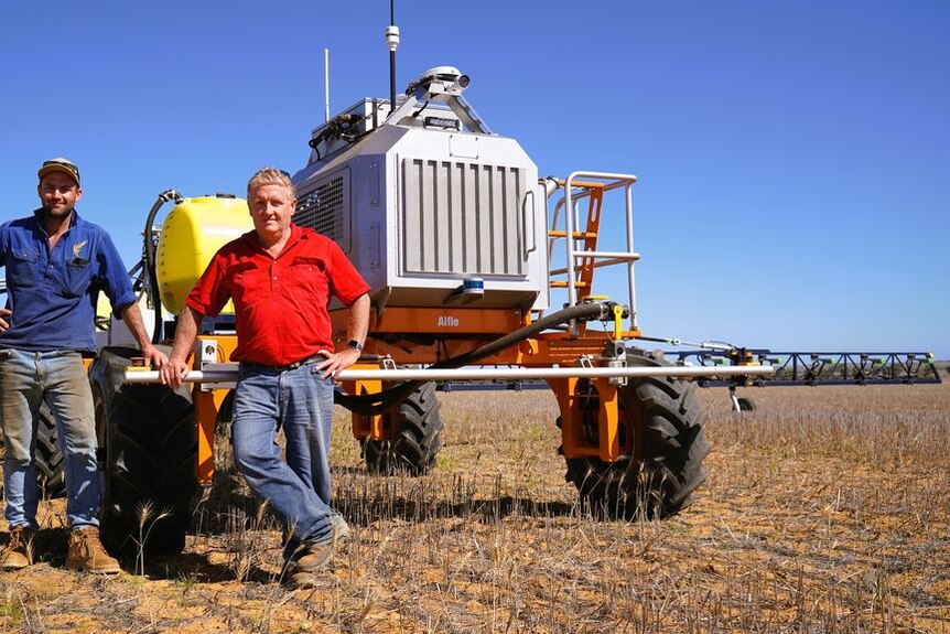 An older and young man stand next to tractor-looking robot in a paddock with stubble, blue skies.