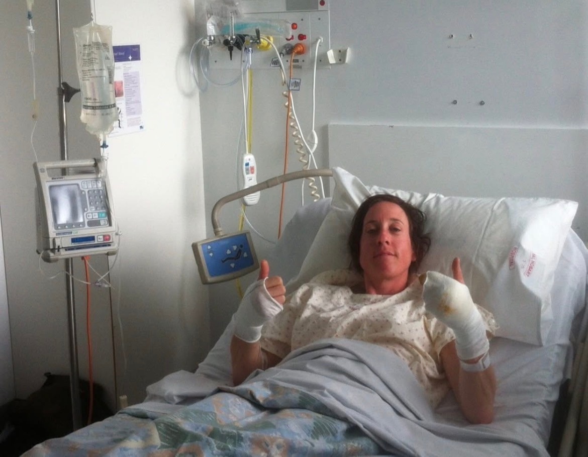 Nicky Stewart holds two thumbs up to the camera while lying in a white hospital bed.