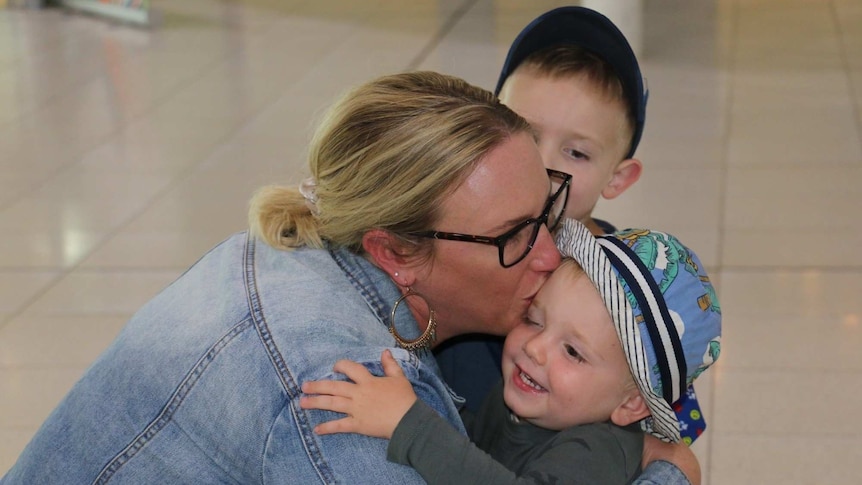 A close up of a woman in an airport terminal bending down and kissing and hugging her young son.