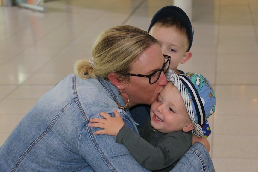 A close up of a woman in an airport terminal bending down and kissing and hugging her young son.