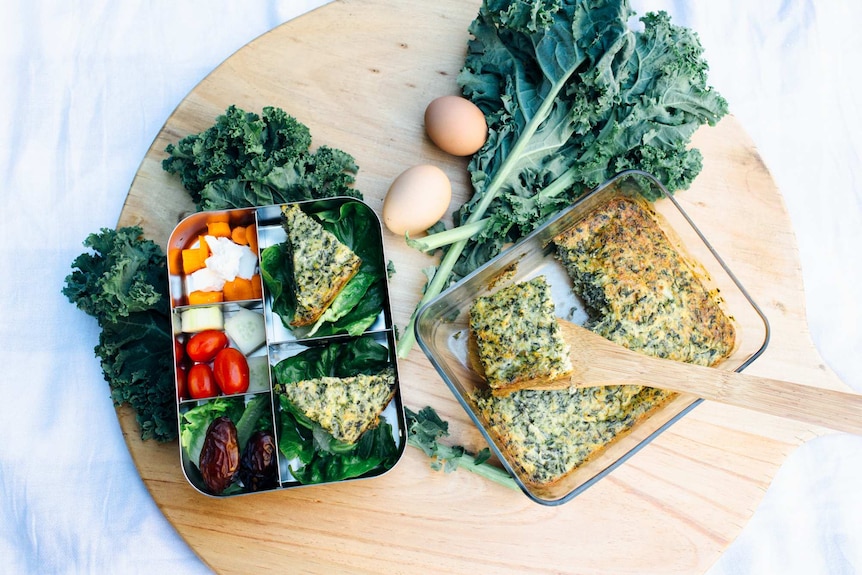 Homemade kale frittata on a chopping board next to a bento box filled with other vegetables and dates.