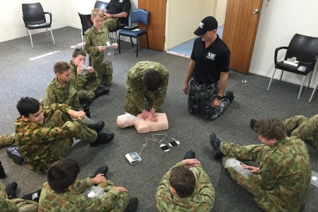 adolescents sit on the floor of a room while an instructor does CPR on a mannequin.