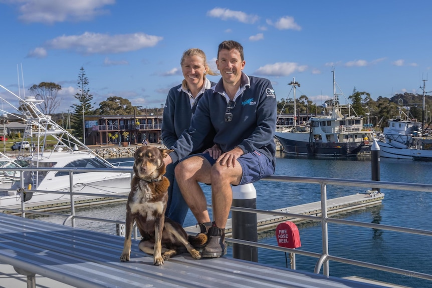 a man and woman sit on the railing of their boat with their dog sitting in front of them