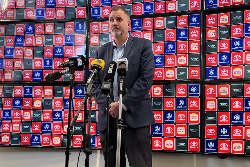 A man wearing a suit speaks to microphones in front of a blue and red banner