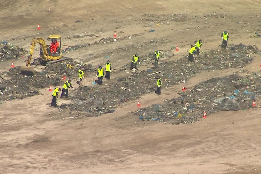 Police in yellow on foot hi-vis vests and an excavator search through piles of rubbish. 