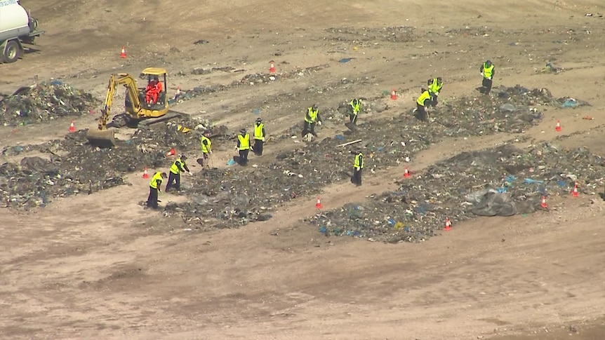 Police in yellow on foot hi-vis vests and an excavator search through piles of rubbish. 