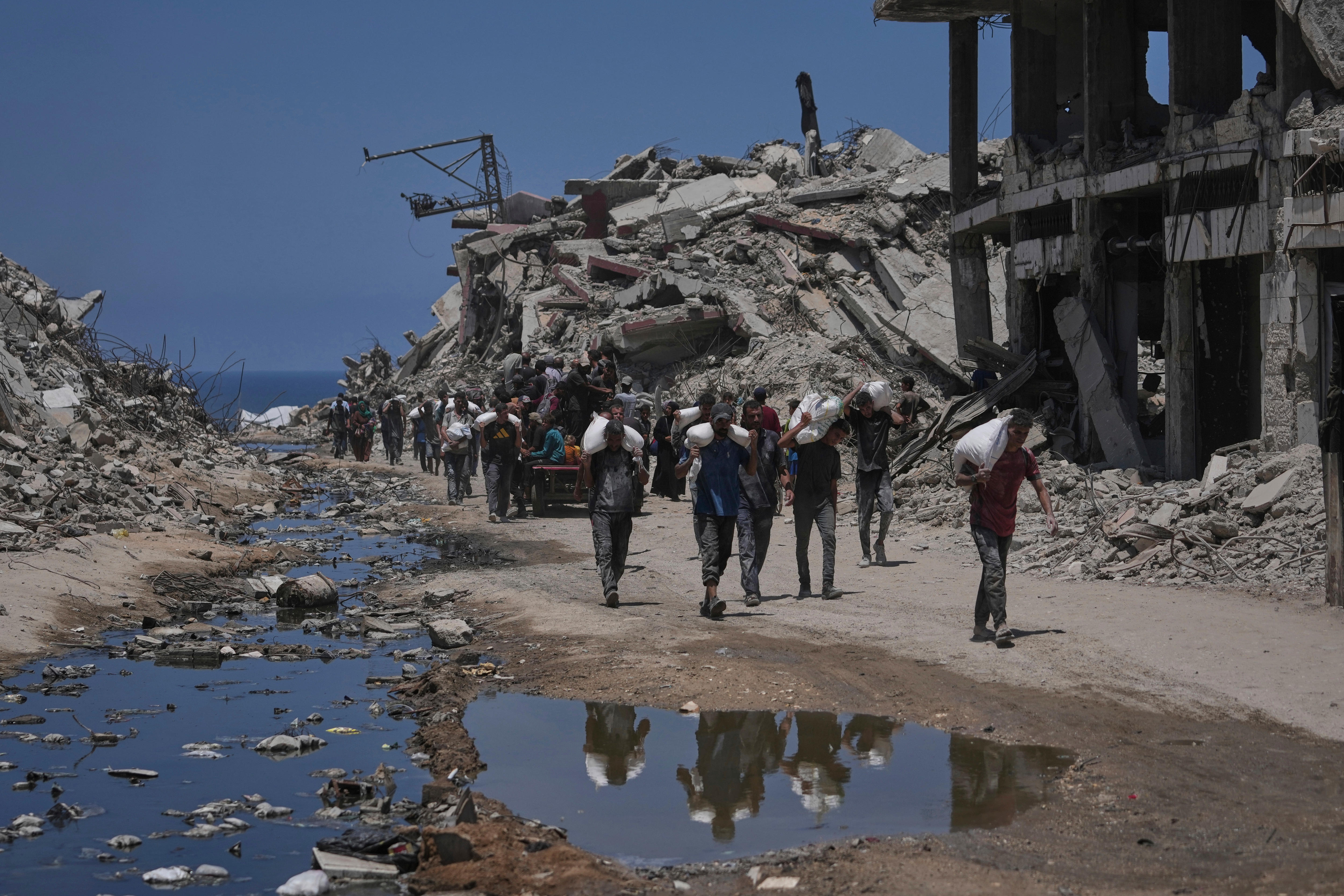 Palestinians carry sacks of flour on the shoulders as they walk down an unseald road past the wreckage of bombed buildings.