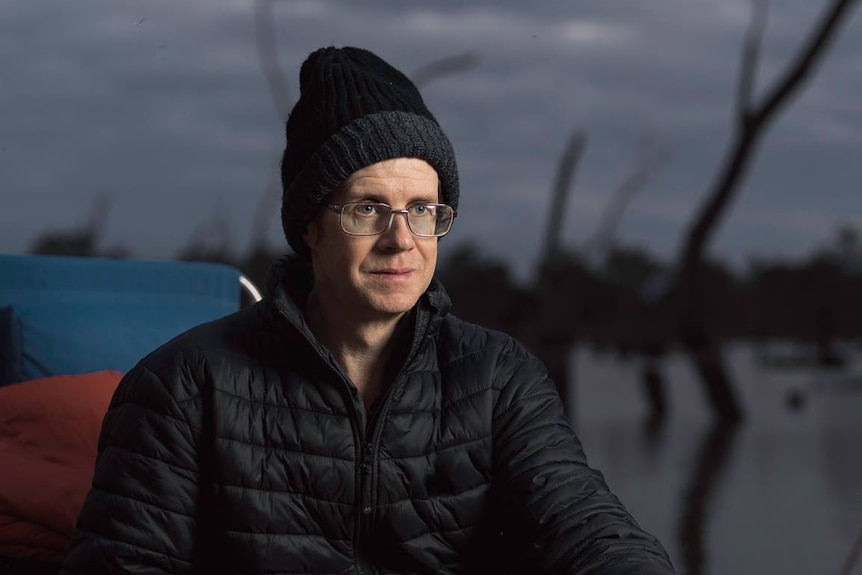 A man wearing a beanie and glasses sits outside next to a lake in the early morning light.
