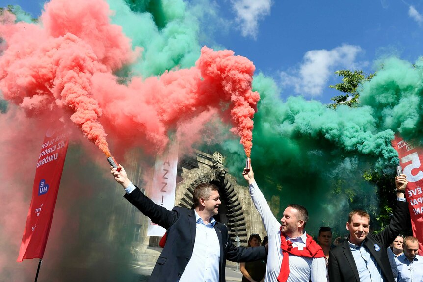 Three men hold flares spouting green and red smoke rising against a blue sky.