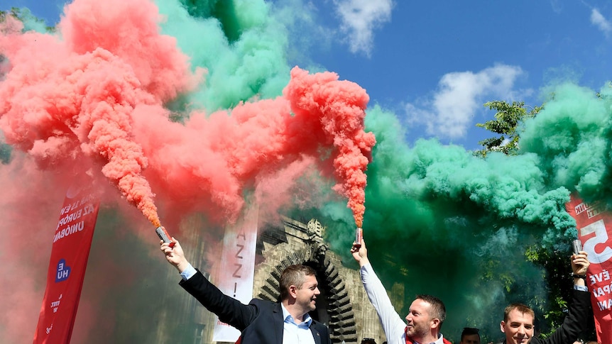 Three men hold flares spouting green and red smoke rising against a blue sky.