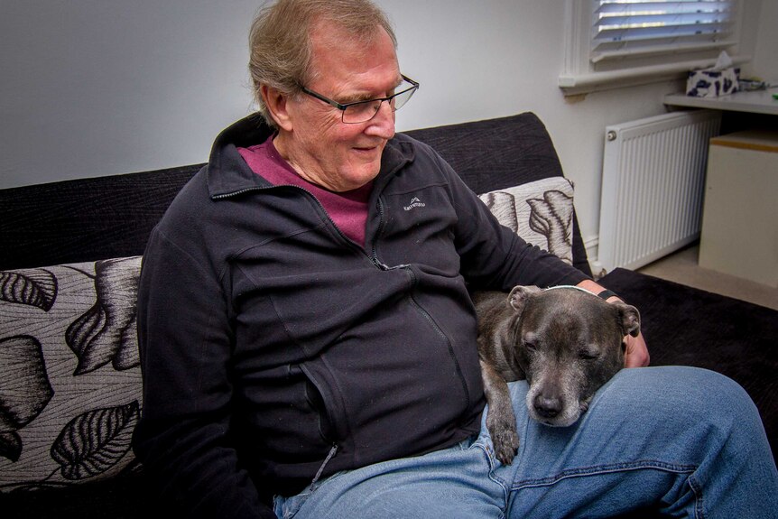 Graeme, dressed in a black jumper, sits on the couch with his dog, Bruiser.