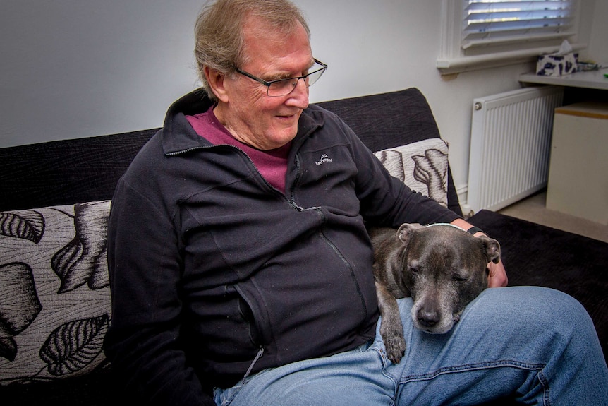 Graeme, dressed in a black jumper, sits on the couch with his dog, Bruiser.