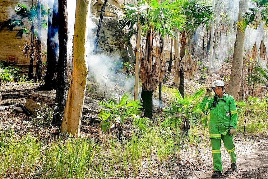 Sheridan Lawton wearing green parks firefighting outfit, burning trees, smoke and greenery behind.