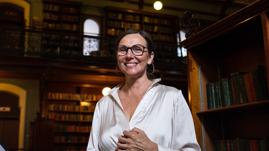 A middle-aged woman with brown hair and glasses, wearing a white silk shirt, stands in a library, looking down at the camera