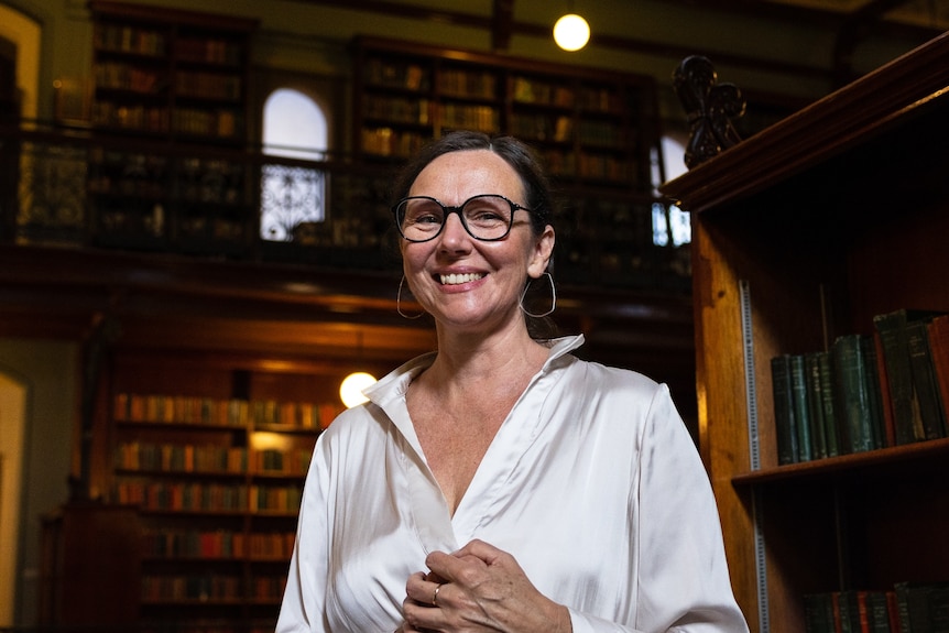 A middle-aged woman with brown hair and glasses, wearing a white silk shirt, stands in a library, looking down at the camera