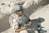 Man sits in military uniform with white sand trench with helmet and gun at knee.