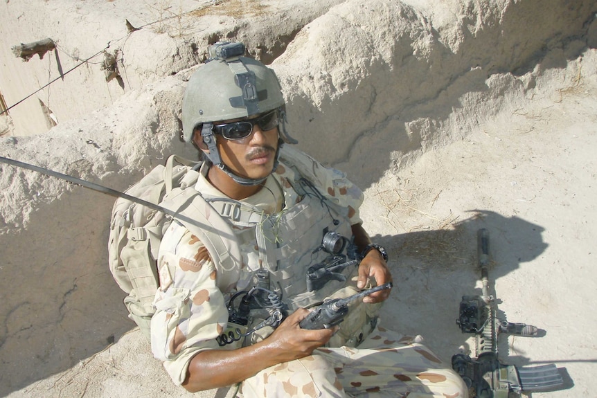 Man sits in military uniform with white sand trench with helmet and gun at knee
