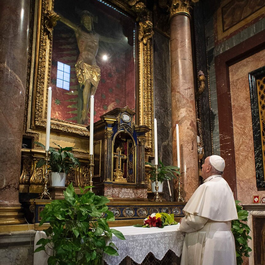 Pope Francis prays in front of a statue of Jesus on a cross that is housed behind glass.