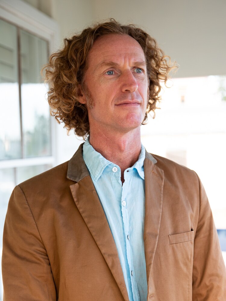 A man with curly hair, wearing a tan jacket.